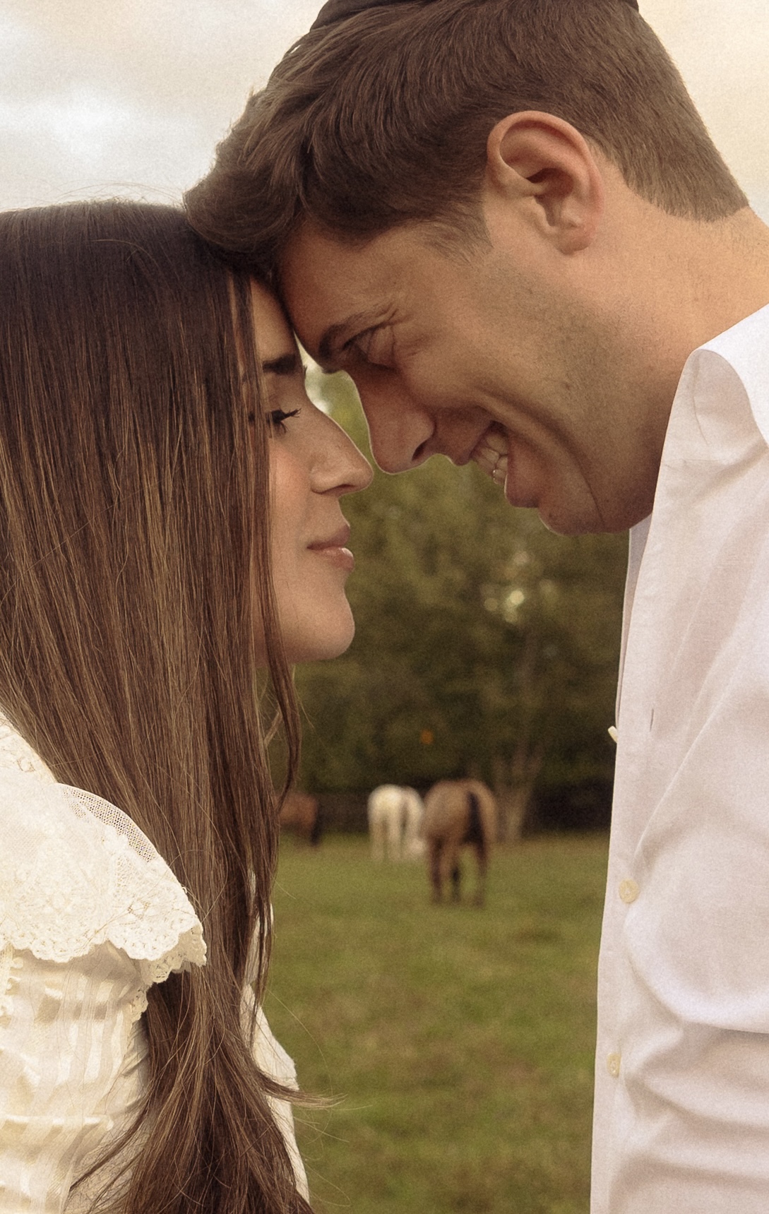 a couple touching foreheads in a green field, a horse grazing behind them